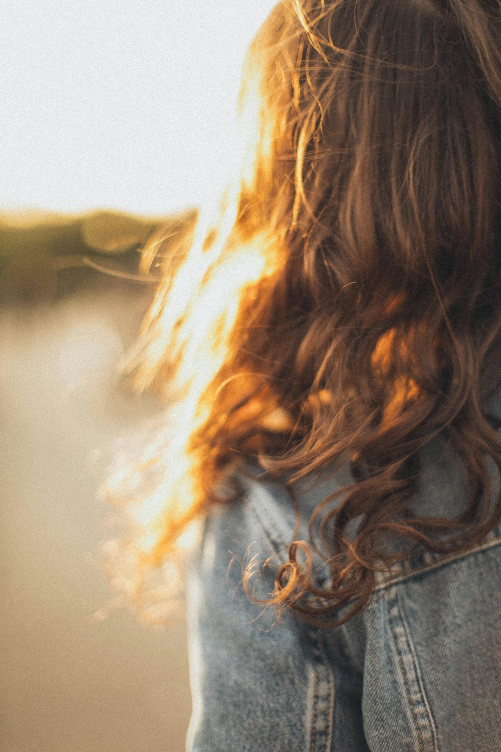 A woman in a denim jacket with long, wavy hair illuminated by sunlight.